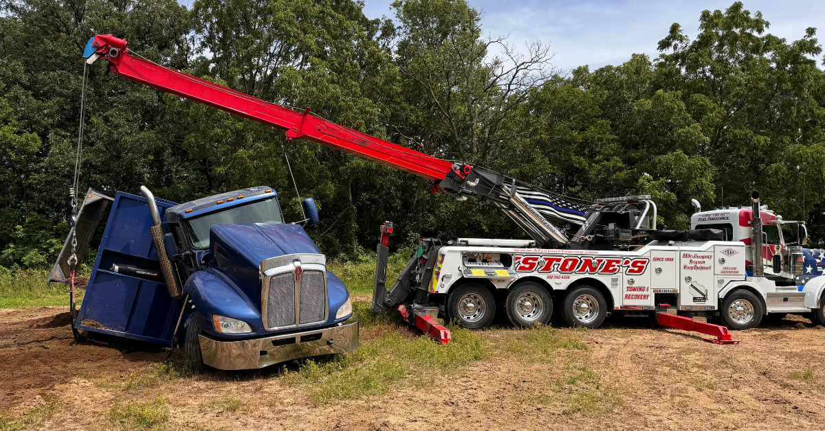 Tow truck recovering a damaged blue semi-truck from a grassy area. The tow truck has a red boom extended.