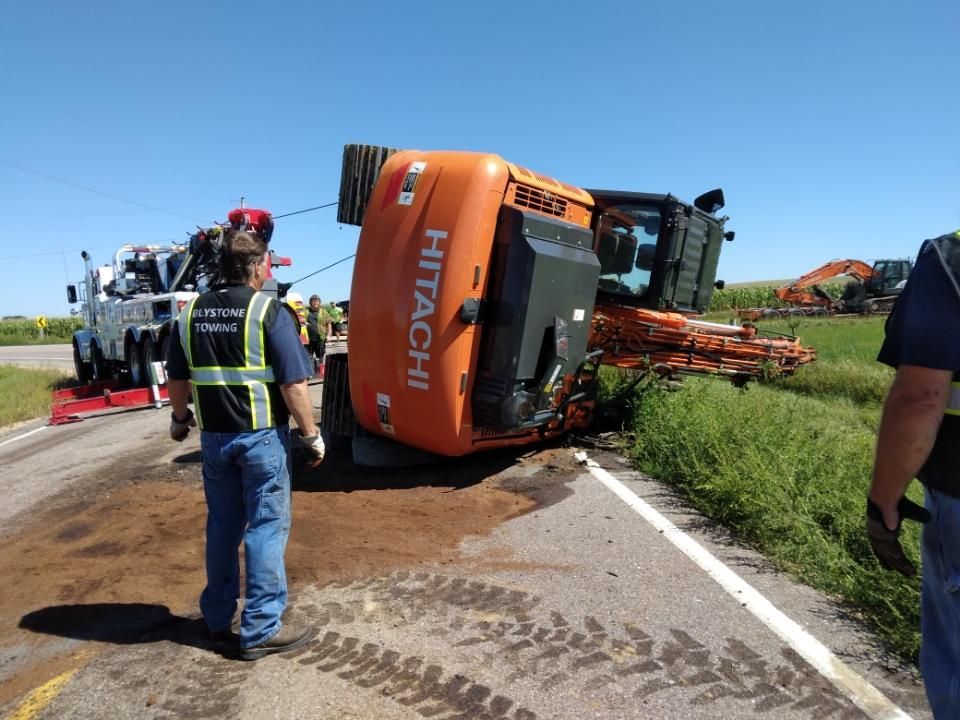 Orange Hitachi construction vehicle overturned on roadside, tow truck and workers present.