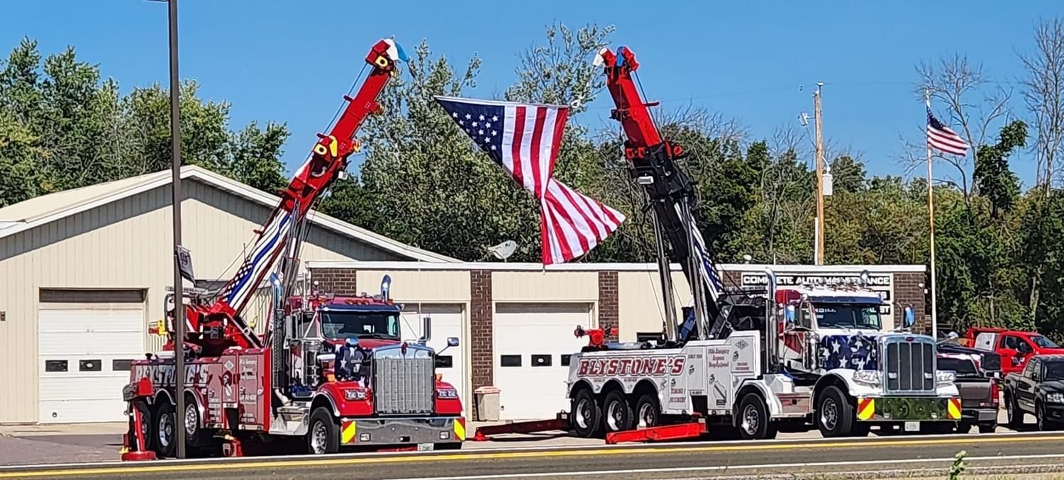 Two tow trucks with raised booms flank an American flag in front of a building.