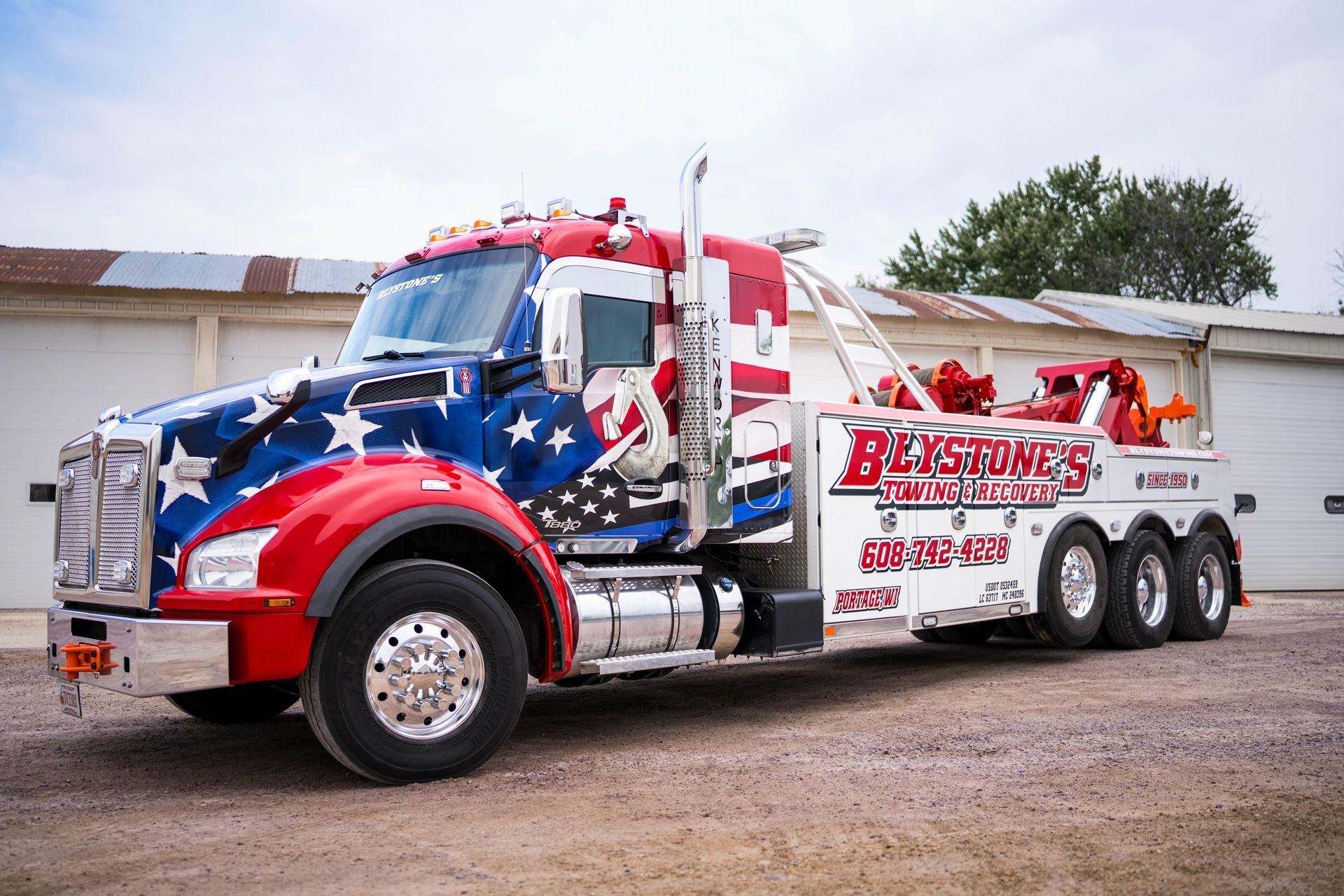 American-flag-themed tow truck parked in front of a white building. Red, white, and blue paint scheme.