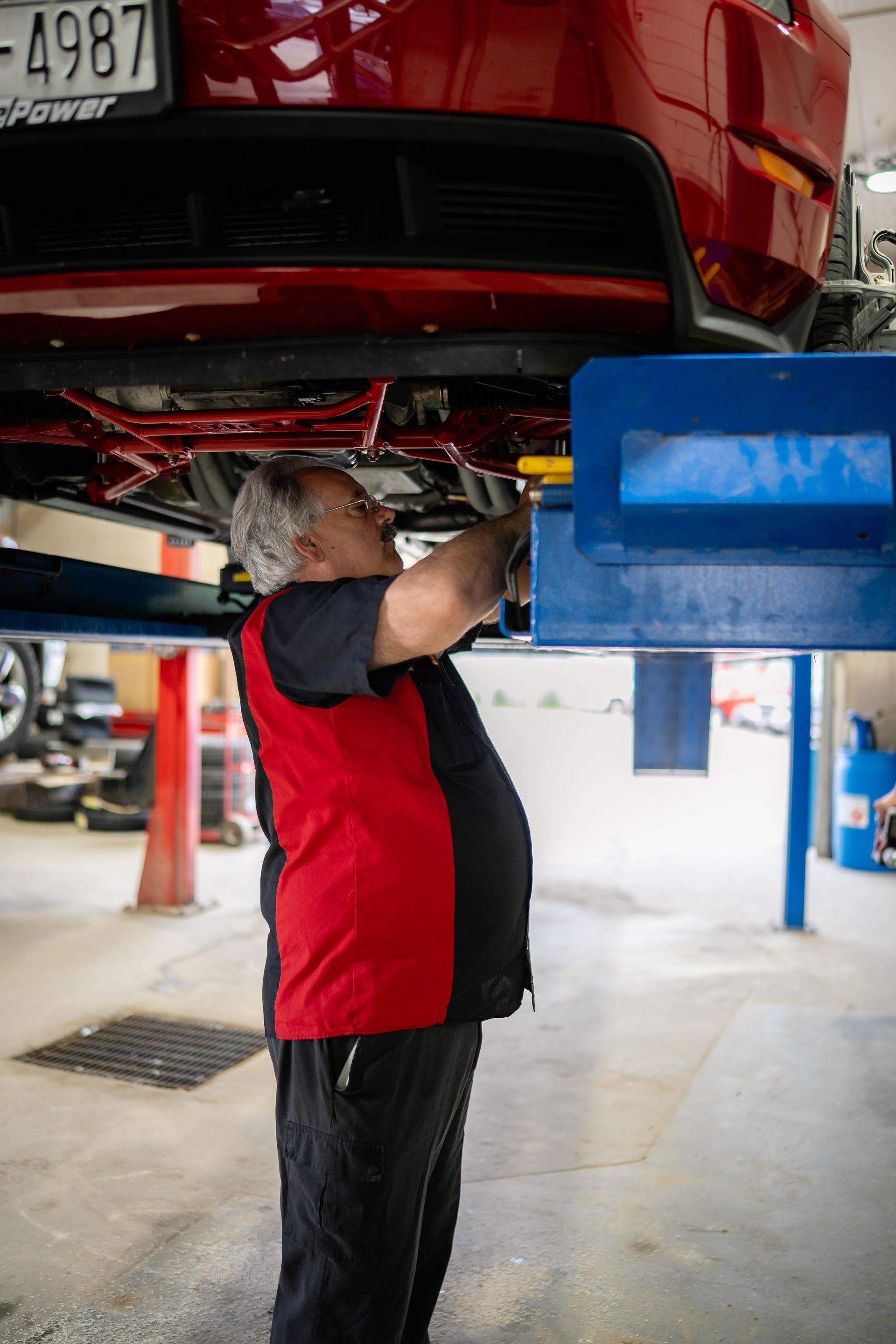 Mechanic working under a red car, on a lift in a garage. Wearing black pants and a red vest.