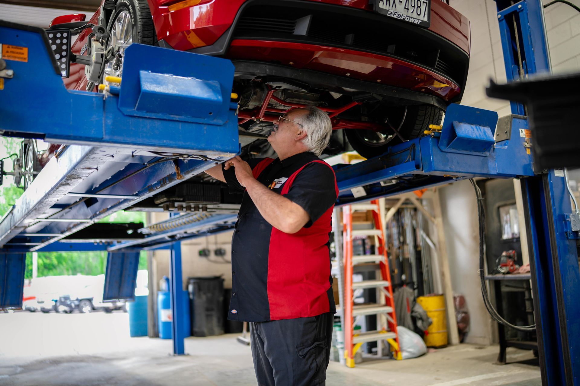 Mechanic inspecting underside of red car raised on a blue lift in a garage.