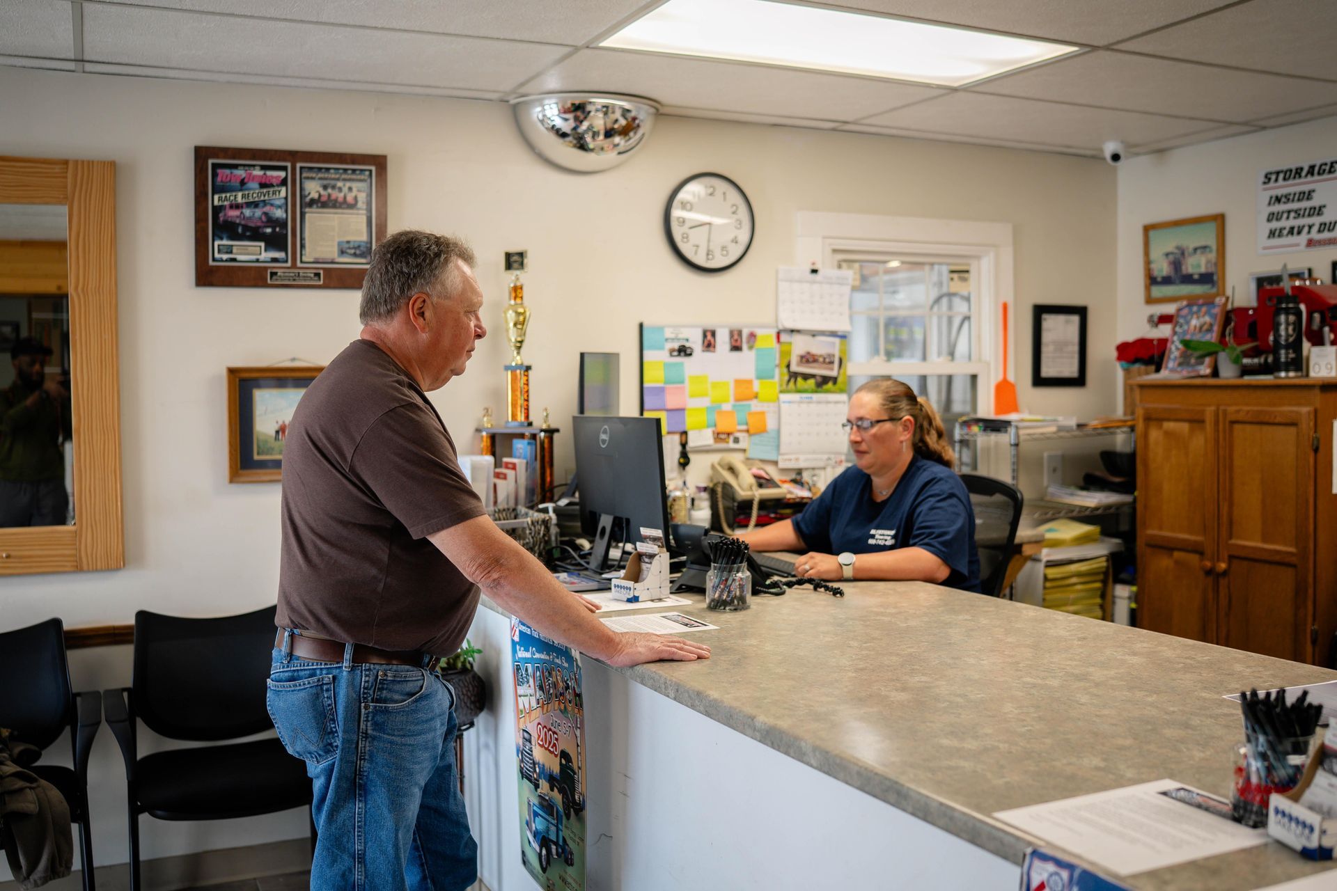 Man talking to a woman at a counter inside a shop. Woman sits at computer. Clock on wall.
