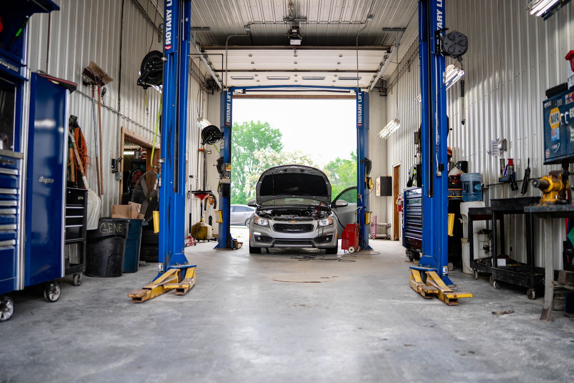 Car in a garage with lift. Open hood, blue lifts, tools, open door to outside.