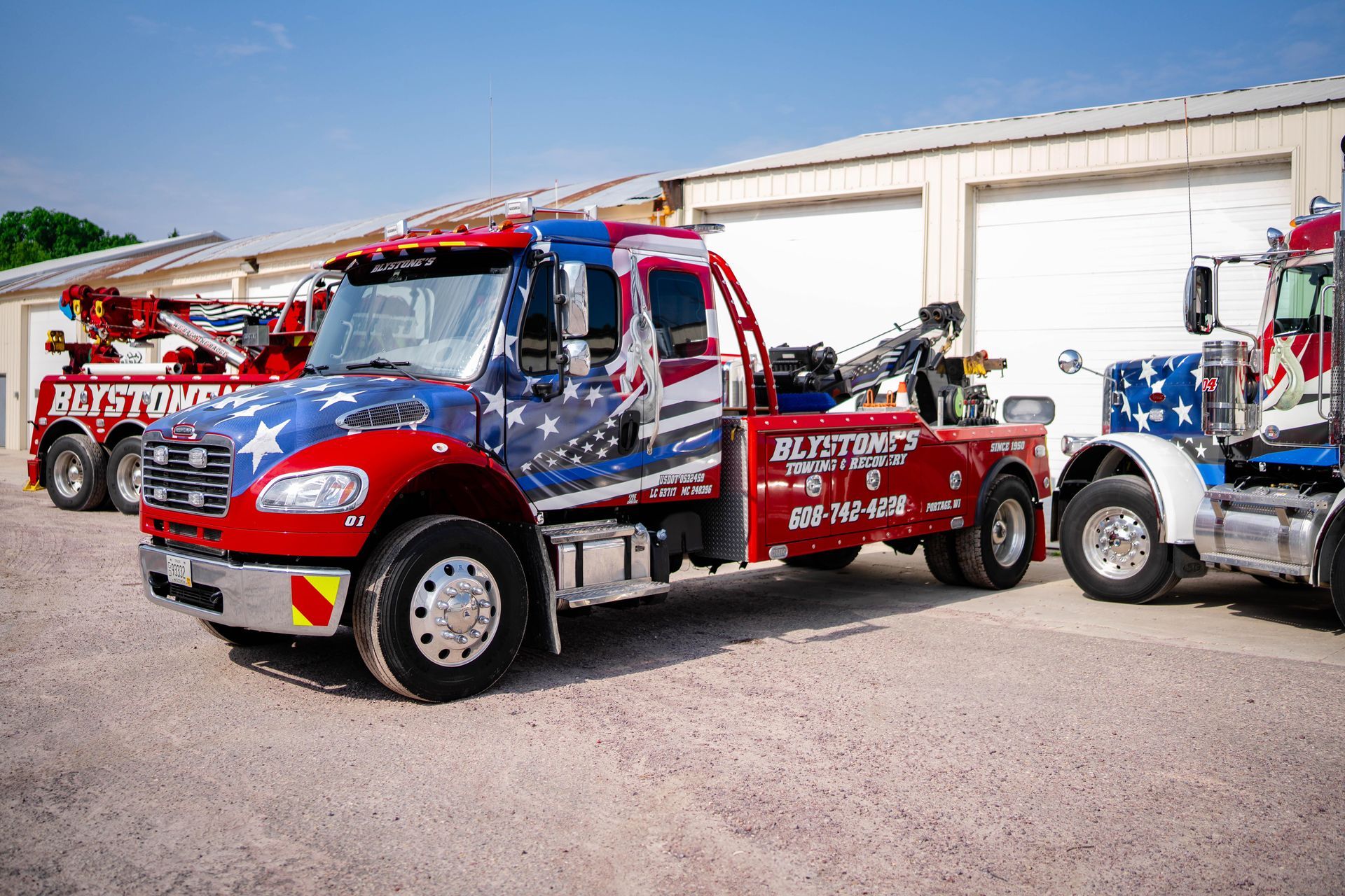 Tow trucks with patriotic paint scheme parked in front of a white building on a sunny day.