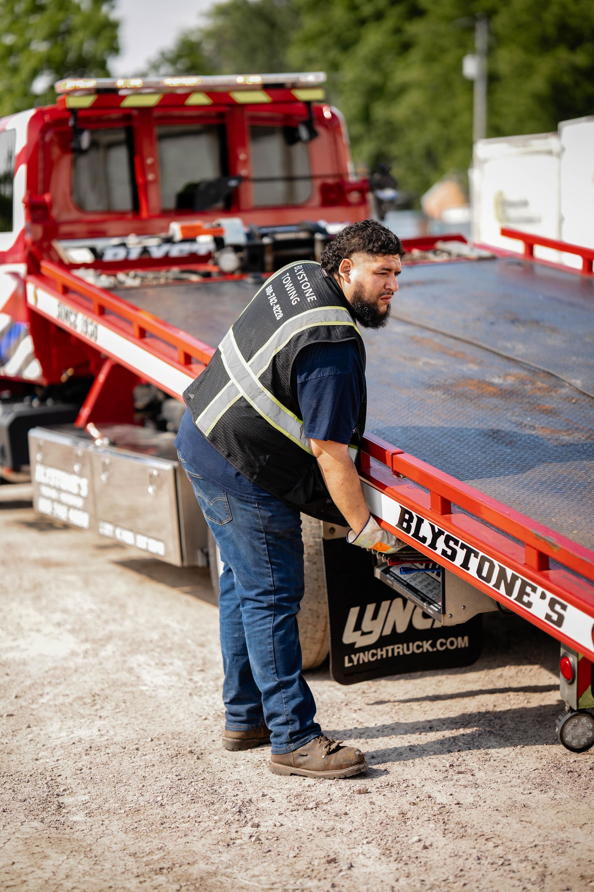 Tow truck operator lowering a flatbed; red truck, gravel lot.