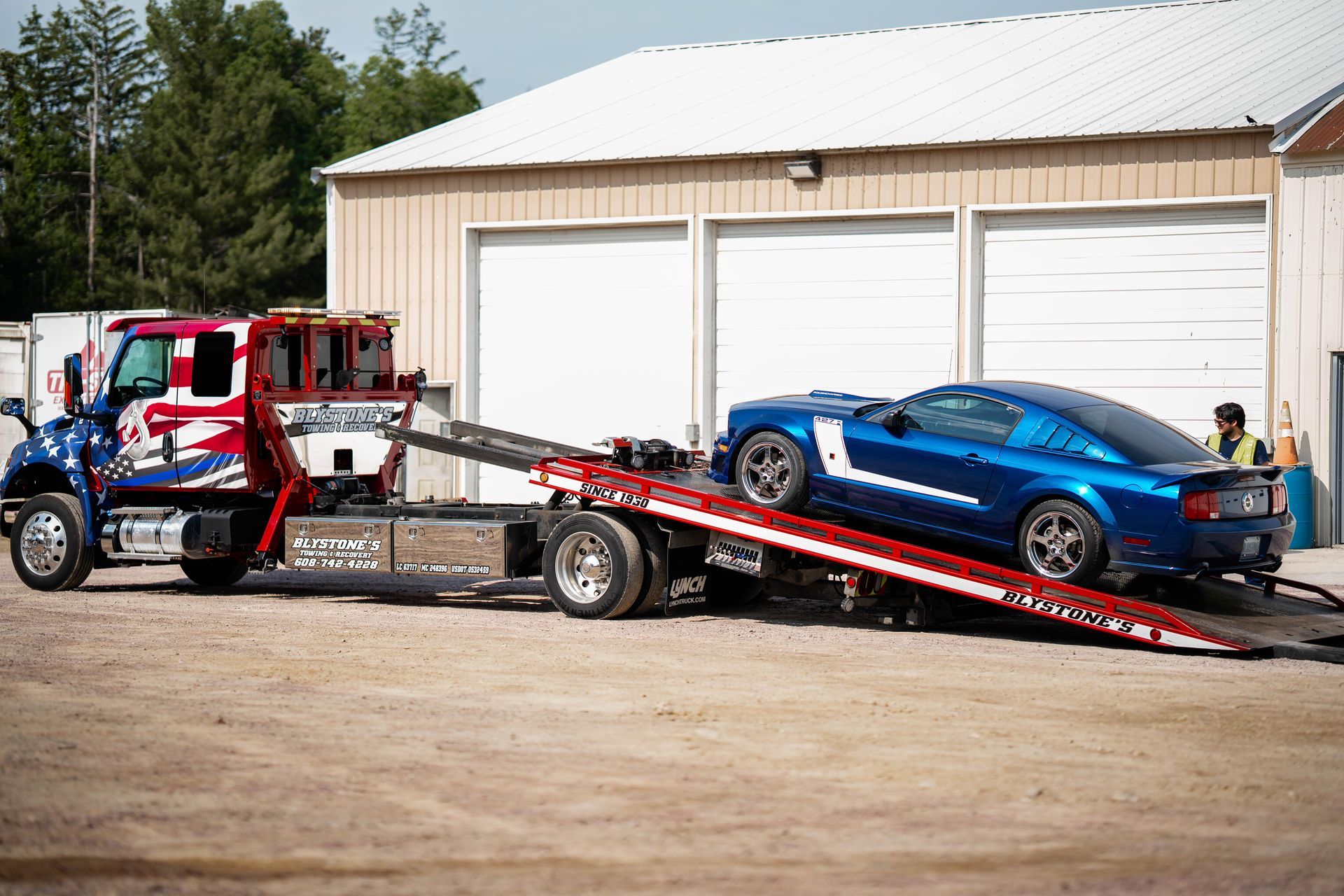 Blue Mustang being loaded onto a red and blue tow truck at a garage.