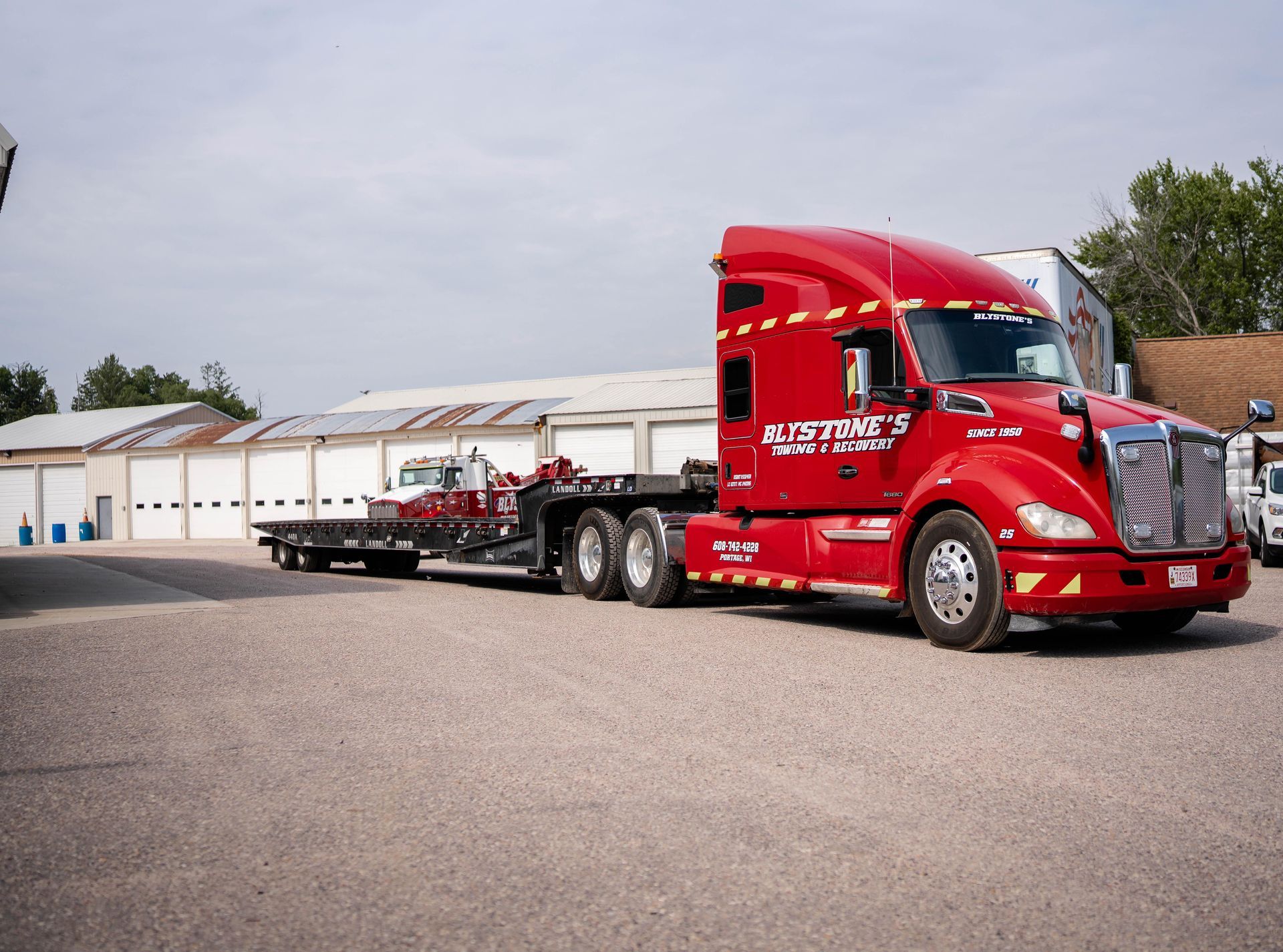 Red semi-truck with flatbed trailer parked on gravel, in front of a building.