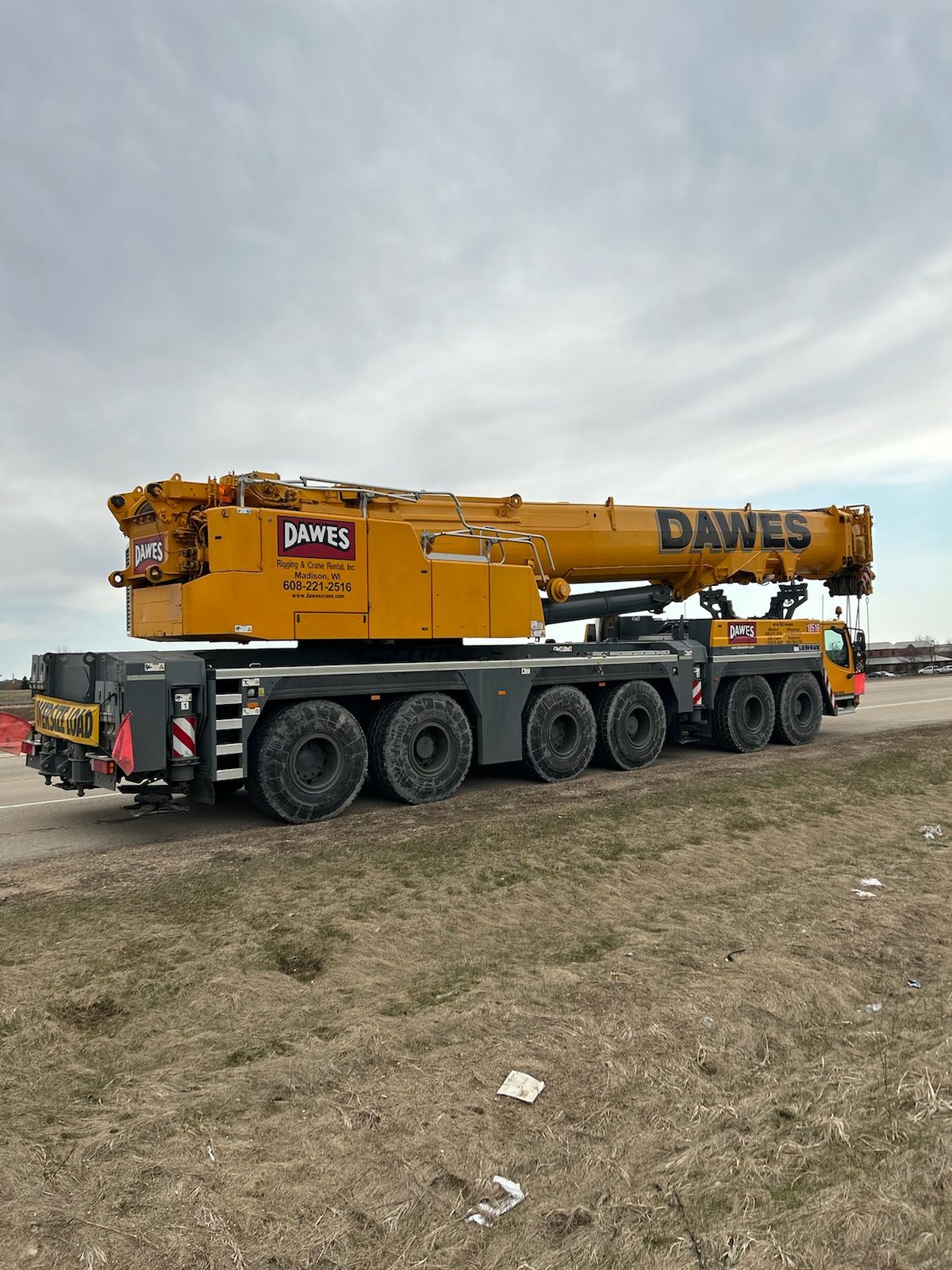 Yellow DAWES crane truck on a flatbed trailer in a grassy lot under a cloudy sky