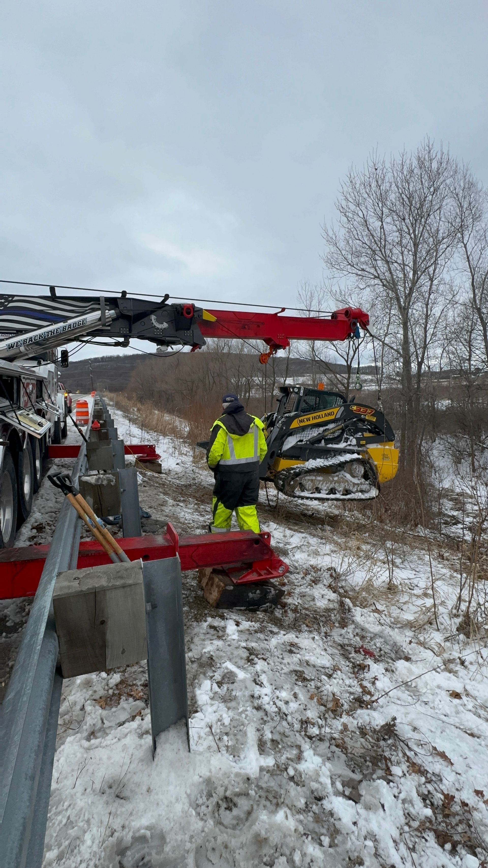 Man in high-vis suit near a yellow skid steer next to a snowy roadside barrier and truck.