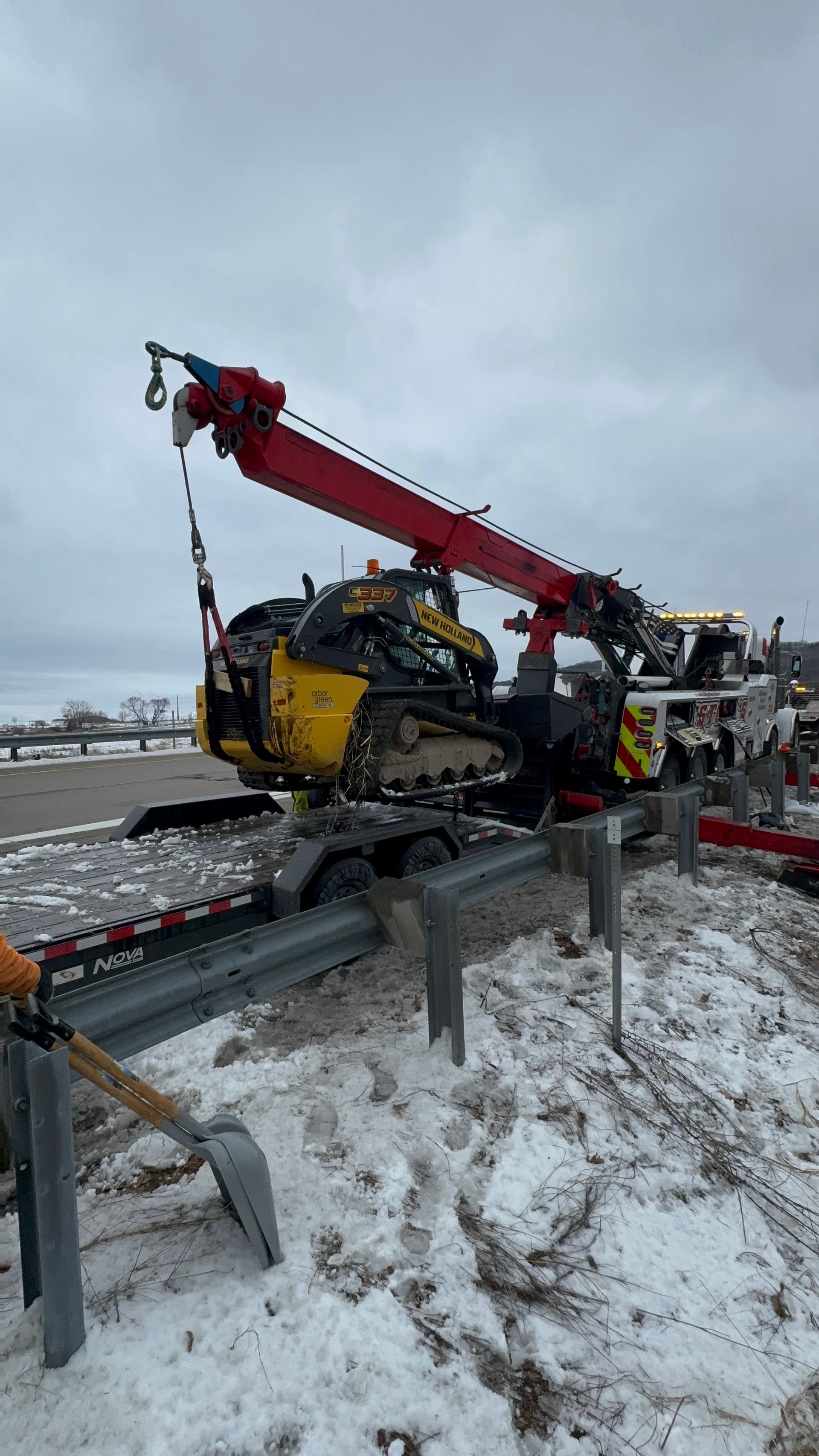 A small yellow excavator being loaded onto a flatbed trailer by a red crane on a snowy roadside.