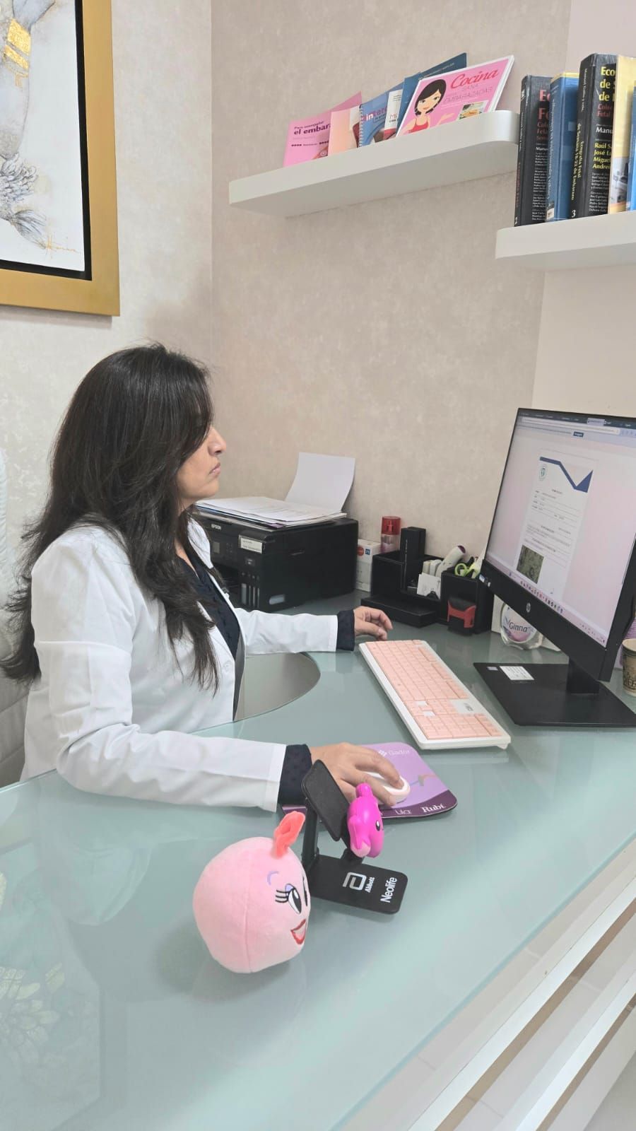 Una mujer con una bata de laboratorio está sentada en un escritorio frente a una computadora.