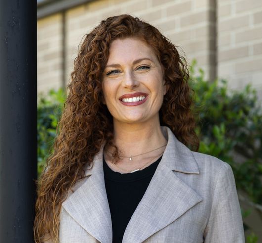 Smiling person with curly hair in a gray coat standing outdoors near a building
