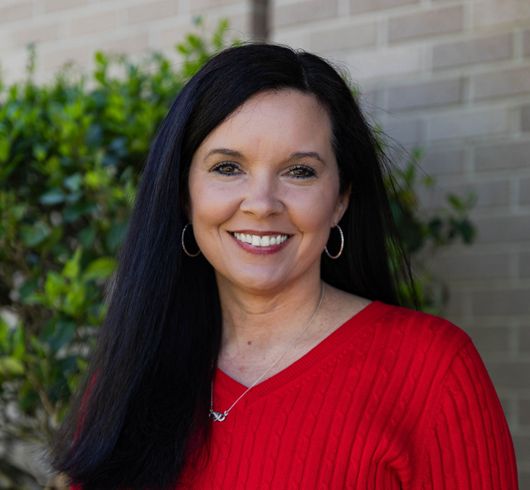 Smiling woman in a red sweater with long dark hair, standing outdoors near greenery.