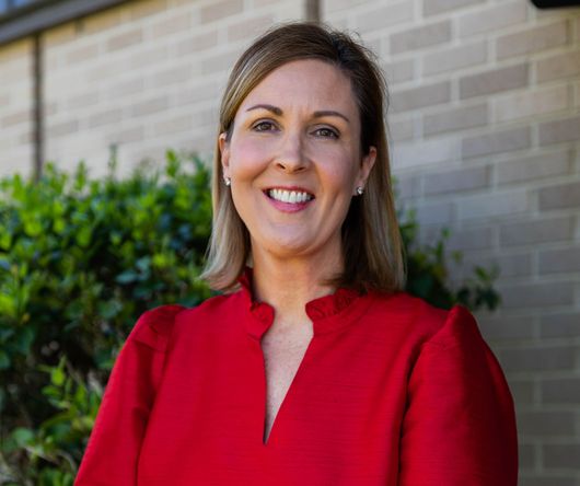 Smiling woman in a red blouse standing outdoors in front of a brick wall and greenery