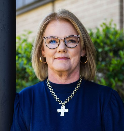 Portrait of a woman wearing glasses, a blue top, and a silver cross necklace outdoors.