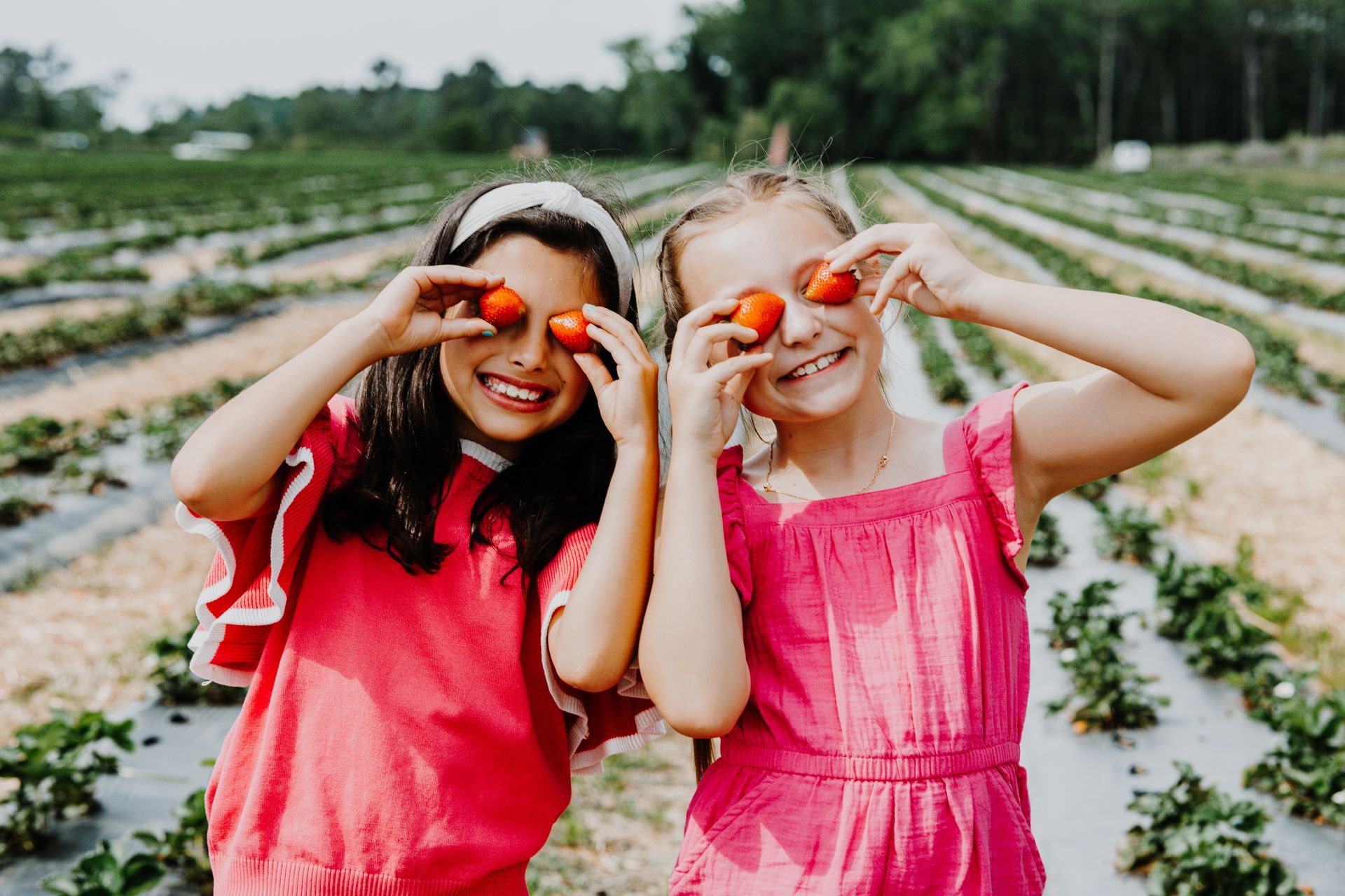 Two children in pink dresses smiling and making heart shapes with their hands in a field