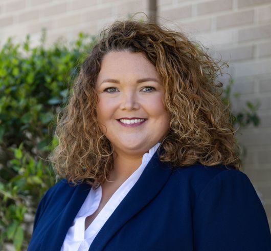 Smiling woman in a navy blazer with curly hair, standing outdoors against greenery.