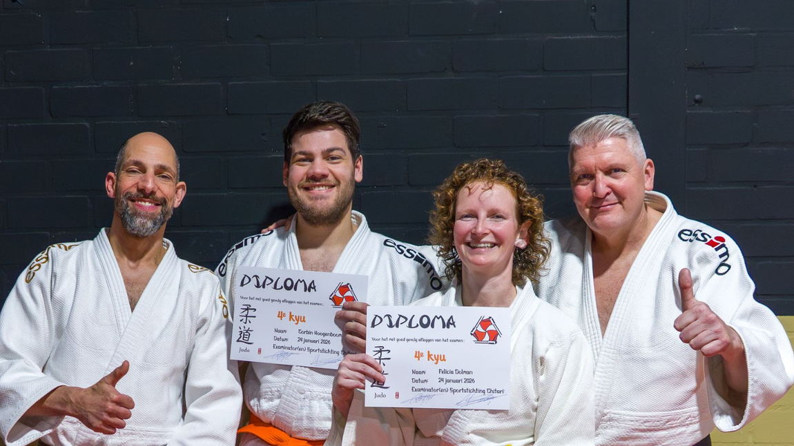 Vier judoka's in witte uniformen glimlachen en houden diploma's vast, waarvan twee hun duim omhoog steken voor een donkere muur.