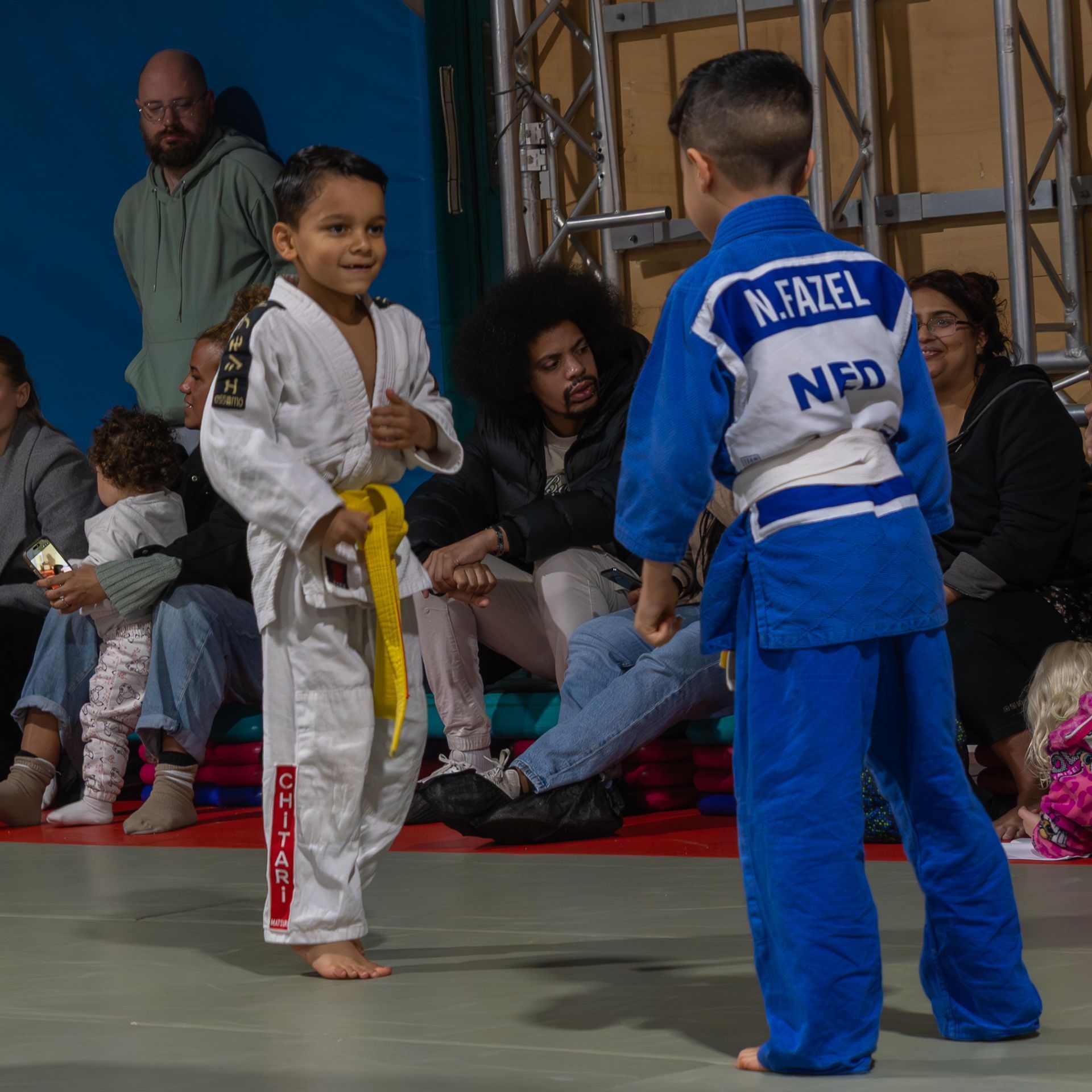 Twee judoka's, de een in een wit judopak met een gele band en de ander in een blauw judopak, staan tegenover elkaar op een mat in een judowedstrijd