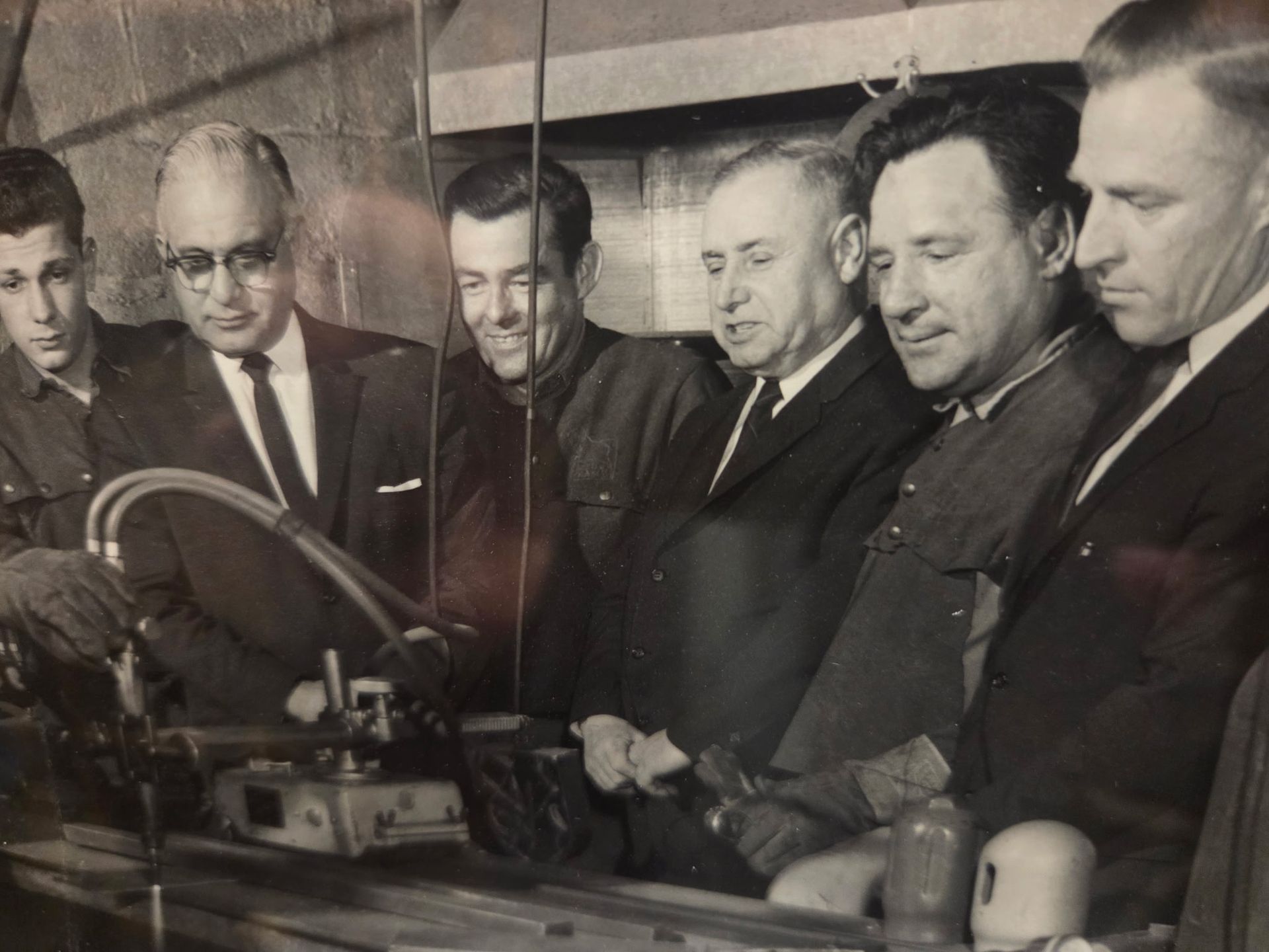 Men gathered around a machine in a workshop, watching closely as one man operates controls.