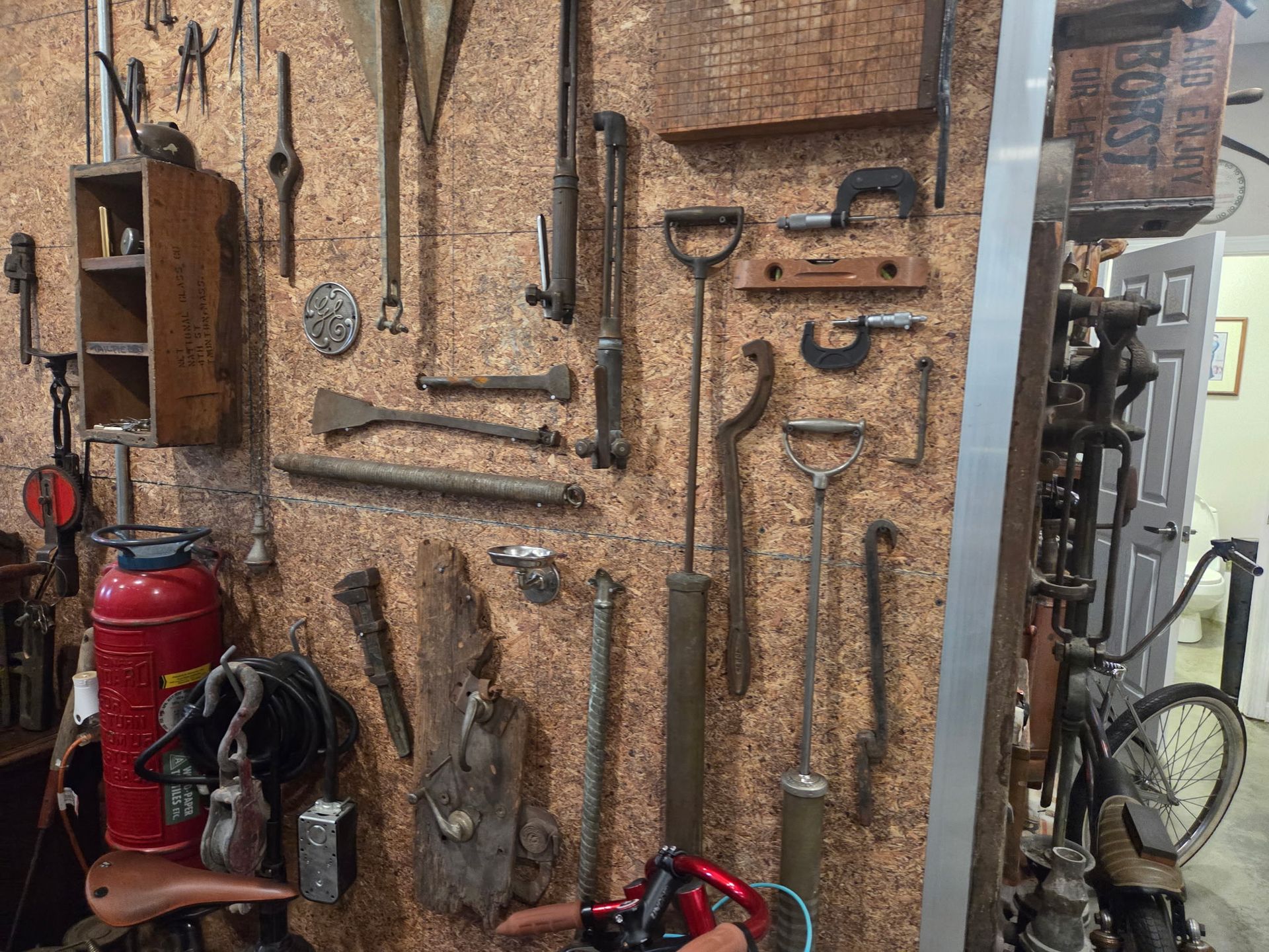 Wall covered with vintage hand tools, with a red fire extinguisher and bicycle in a workshop corner