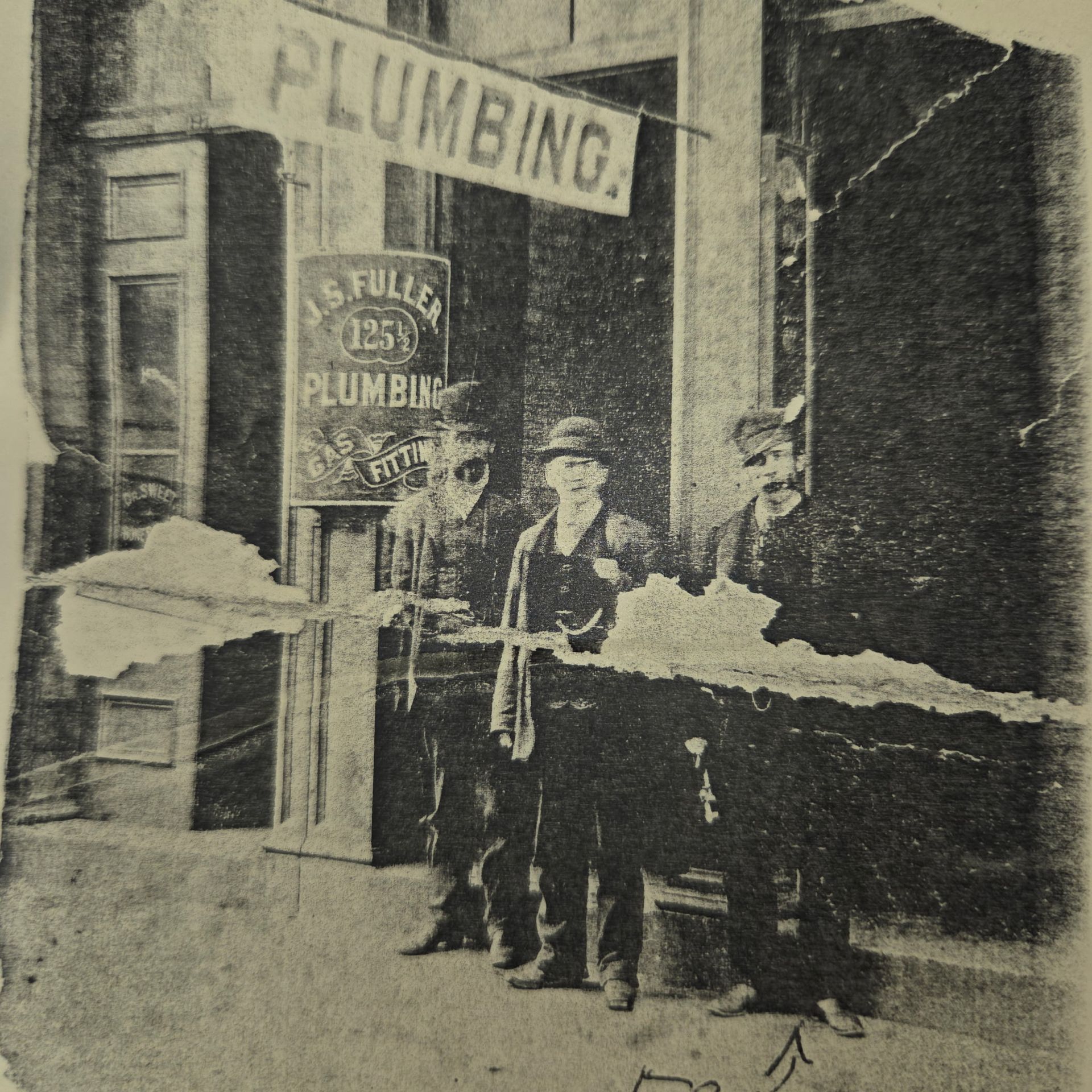 Black-and-white photo of three men outside a plumbing shop with a “PLUMBING” sign.