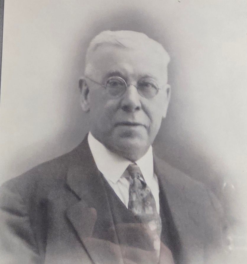 Black-and-white portrait of a man in a suit, tie, and round glasses, facing the camera.
