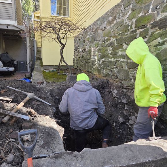 Workers in neon jackets digging a trench beside a stone wall and yellow house.