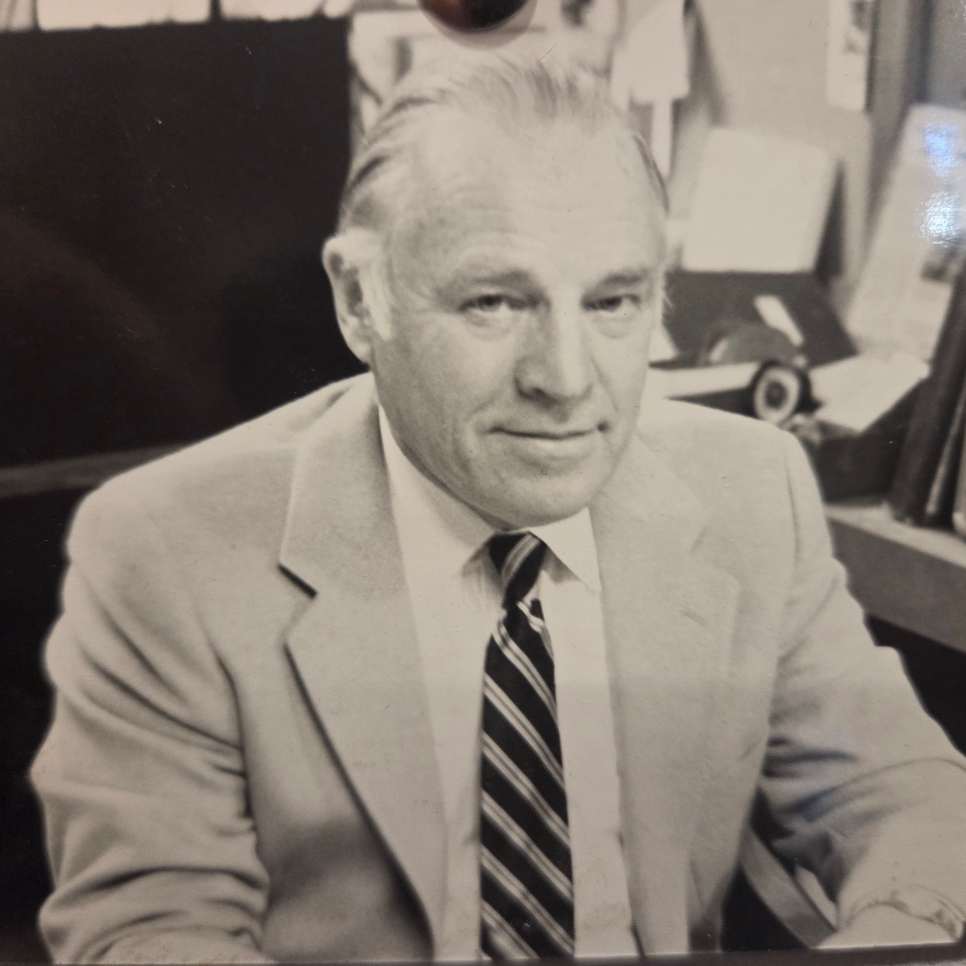Smiling man in a light suit and striped tie seated at a desk in an office.