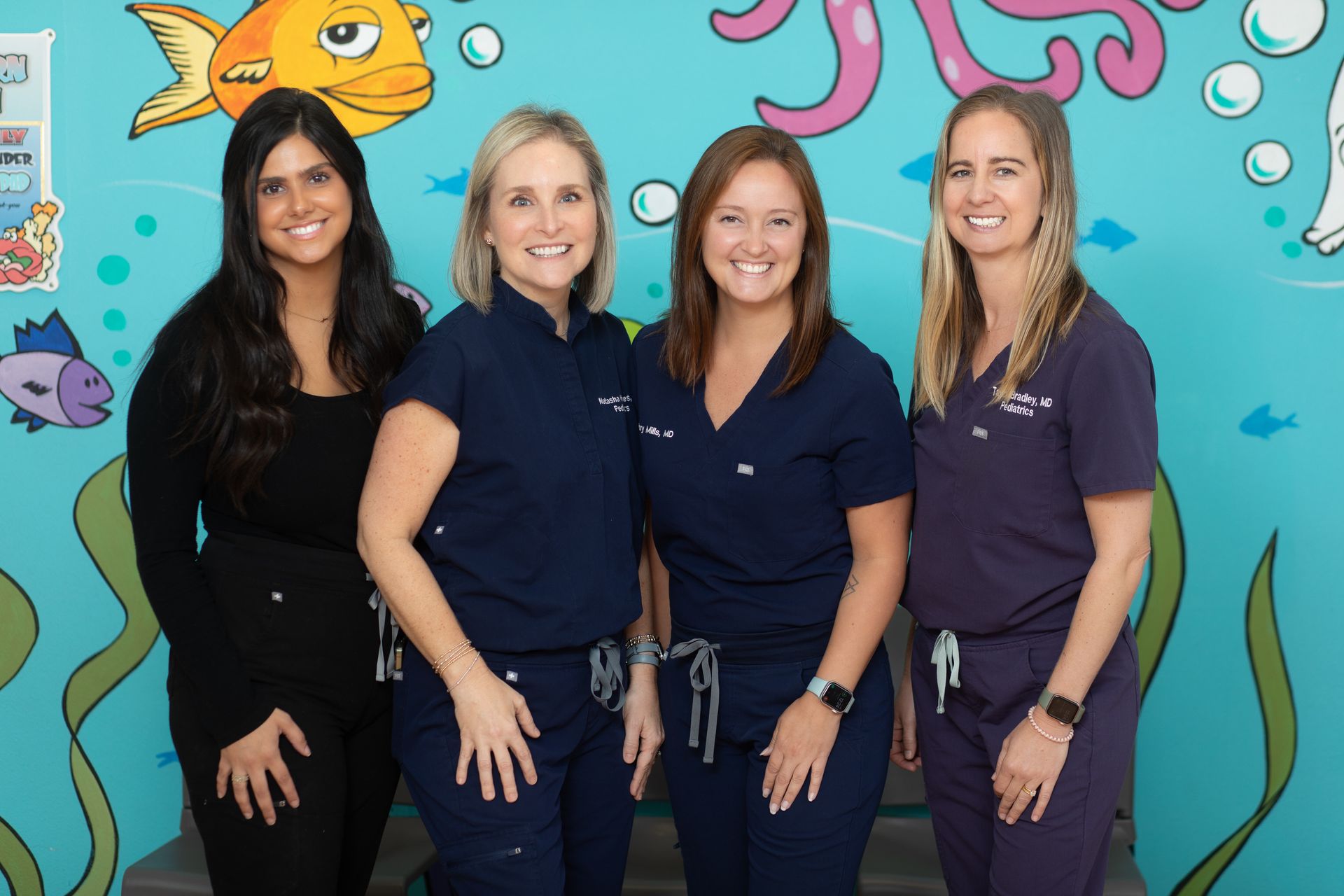 Four women in scrubs are posing for a picture in front of a mural of fish and octopus.