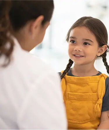 Girl with pigtails smiles at person in white coat; inside a well-lit space.