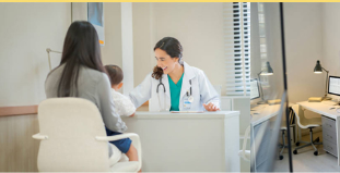 Doctor speaking with a patient in a bright clinic office at a desk with a computer and lamp.