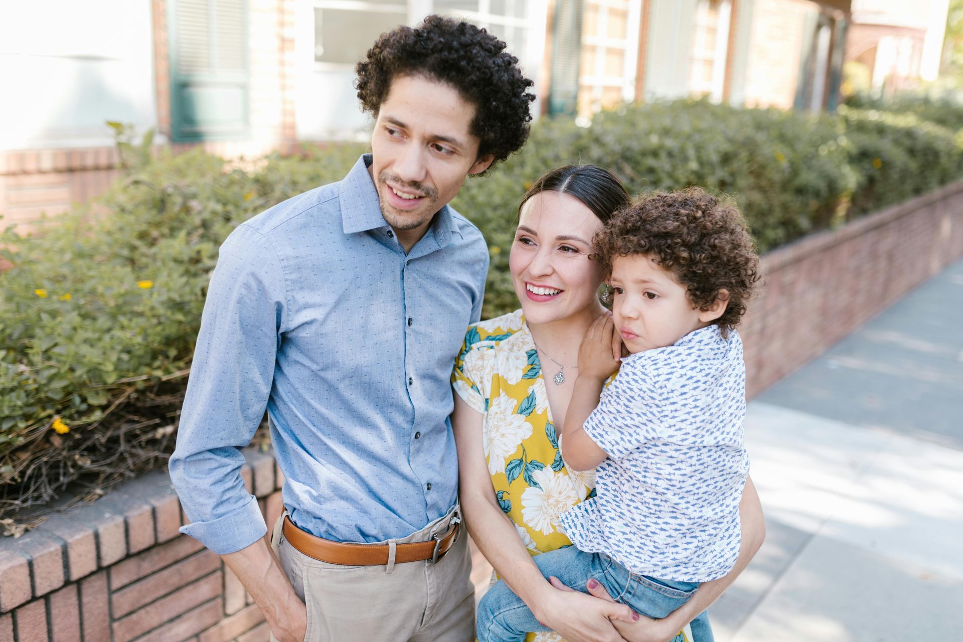 Three people smiling outdoors by a brick wall, with one holding a child.