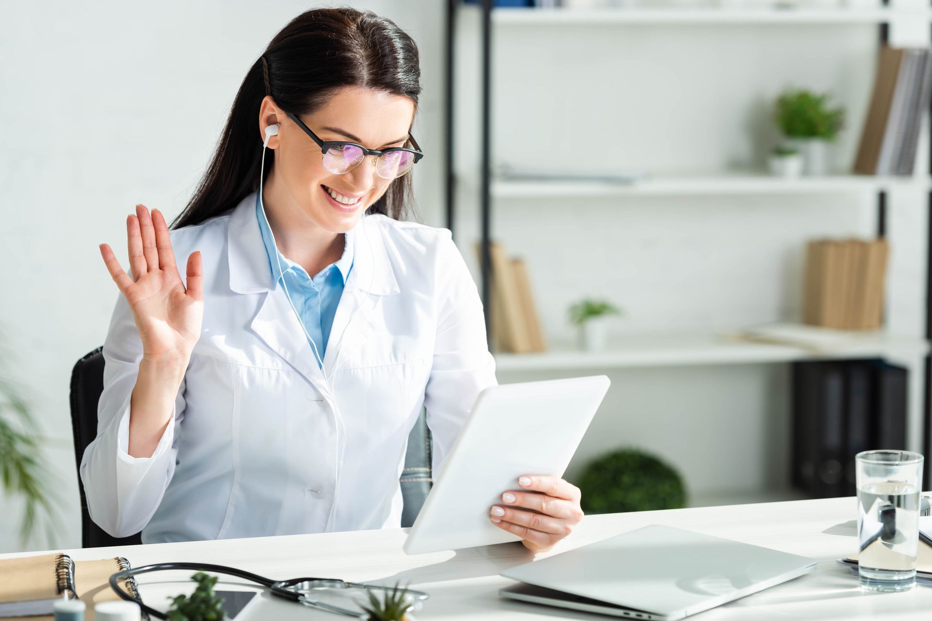 Un médico con bata blanca saluda sonriente a una tableta durante una consulta virtual.