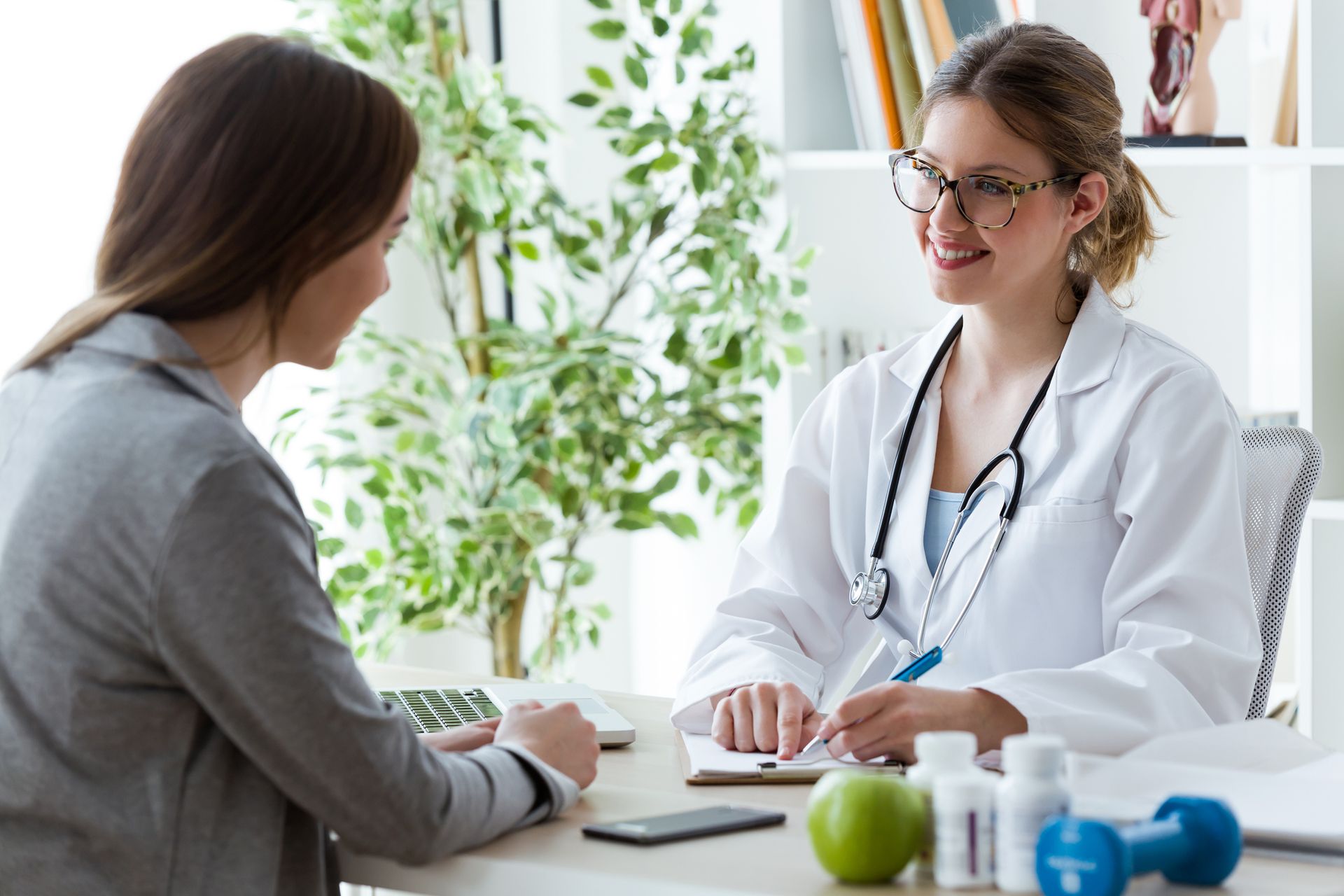 Mujer consultando con un médico en un consultorio; el médico sonríe, sosteniendo un bolígrafo y una libreta.