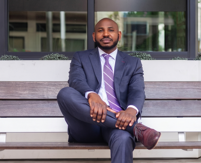 Man in a navy suit sitting cross-legged on a bench in front of a building