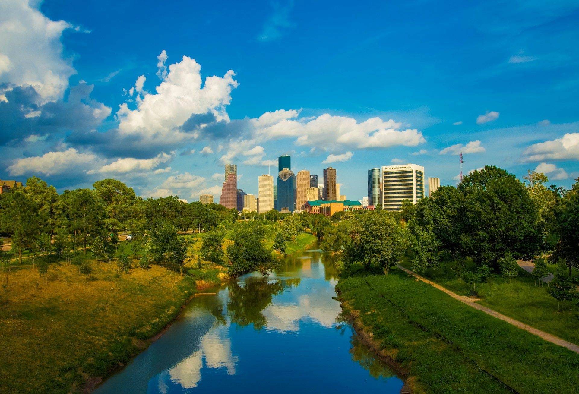City skyline reflected in a calm river, framed by green trees under a bright blue sky with clouds