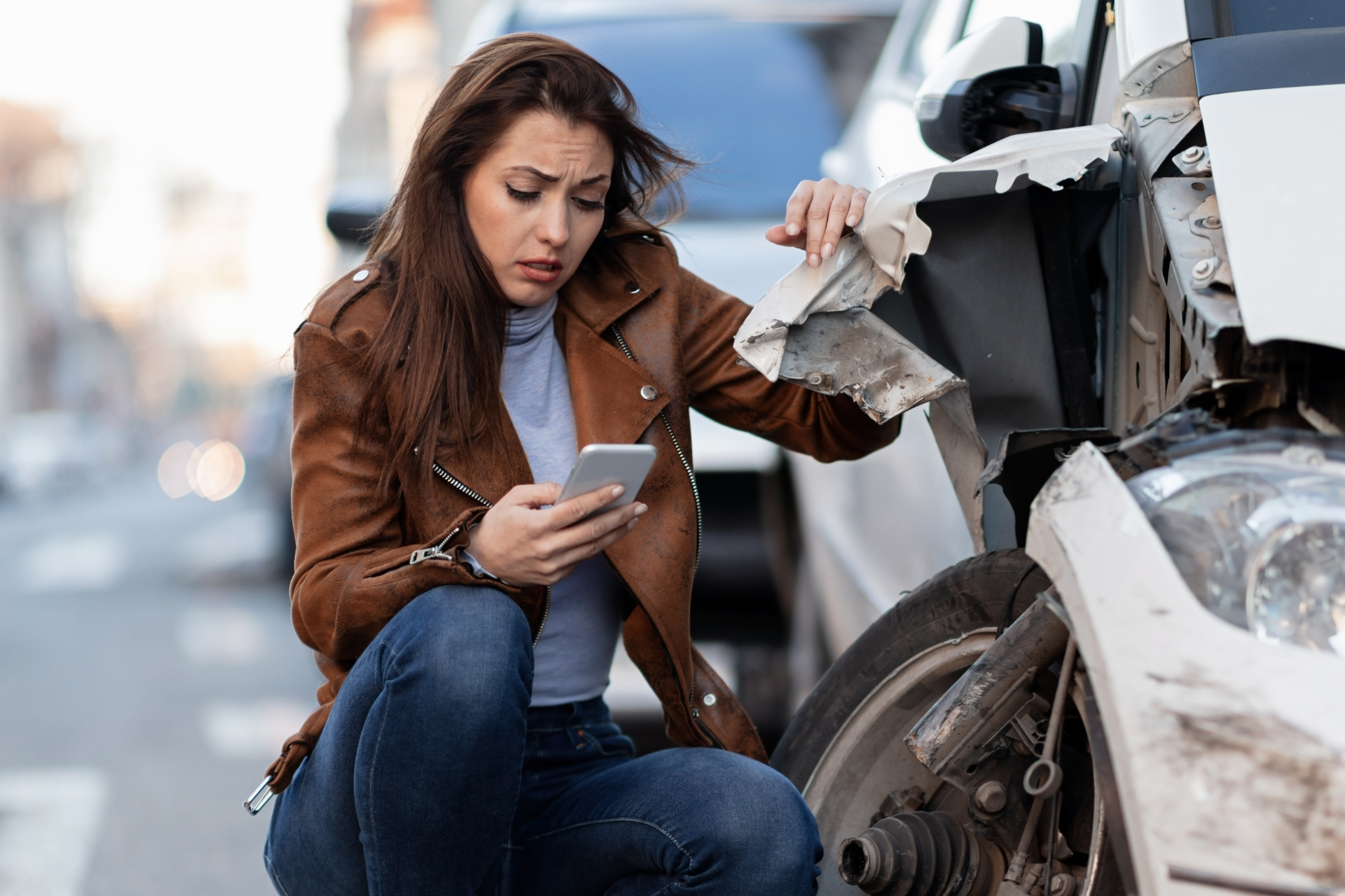 Woman kneels beside a crashed car, looking at her phone with a damaged front bumper in view