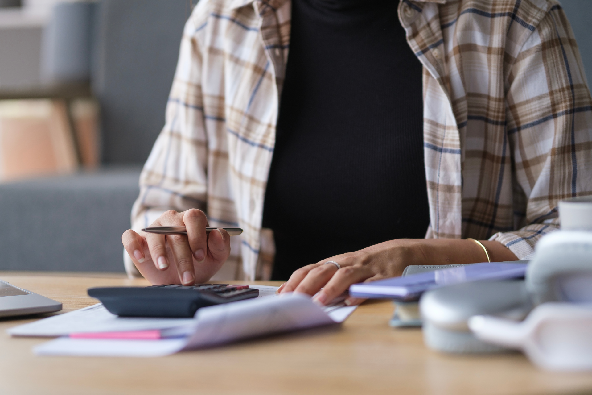 Person writing in a notebook at a desk, wearing a plaid shirt, with papers and a cup nearby.