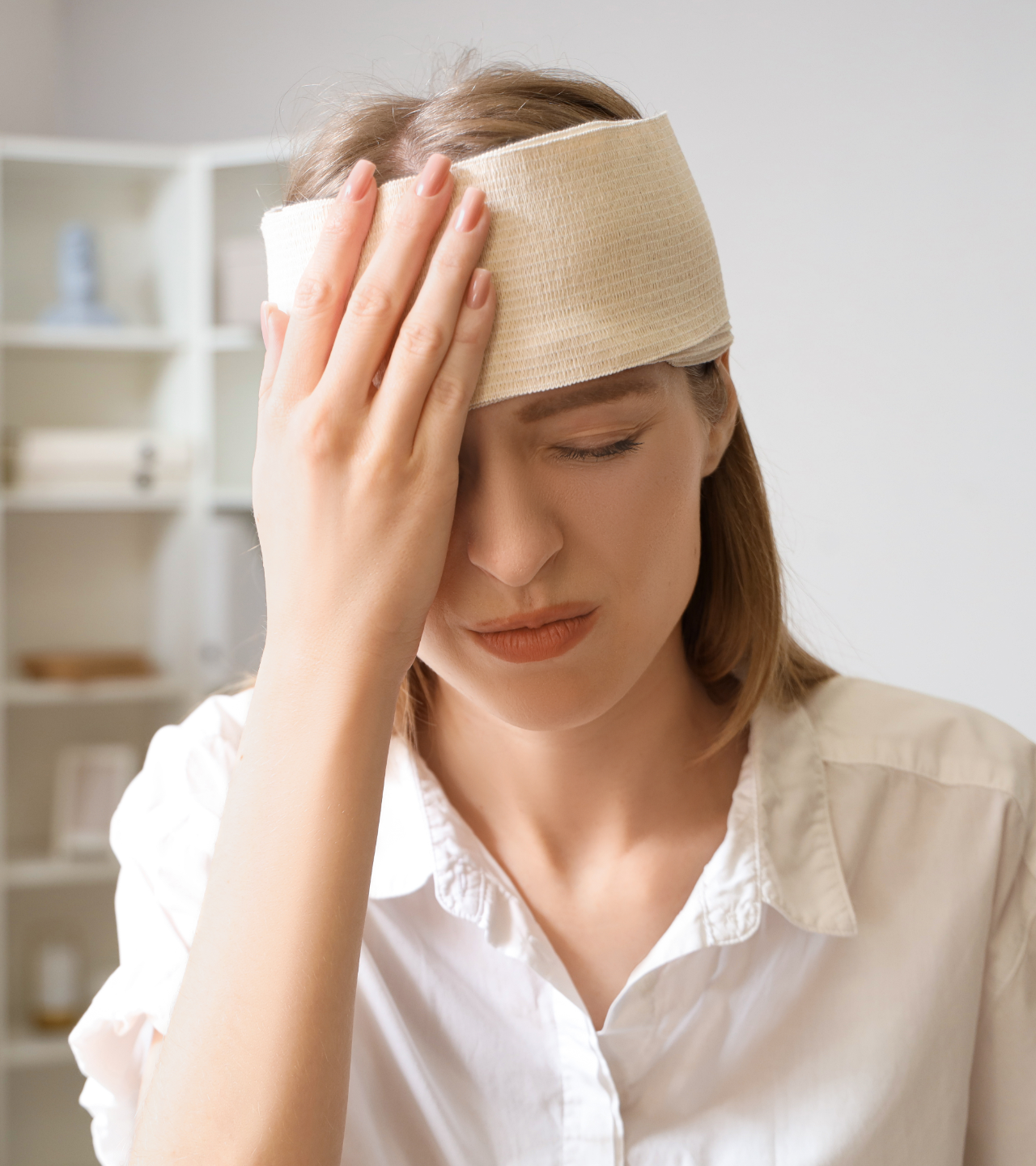 Person with a white bandage on their forehead, holding their head and looking down in discomfort.