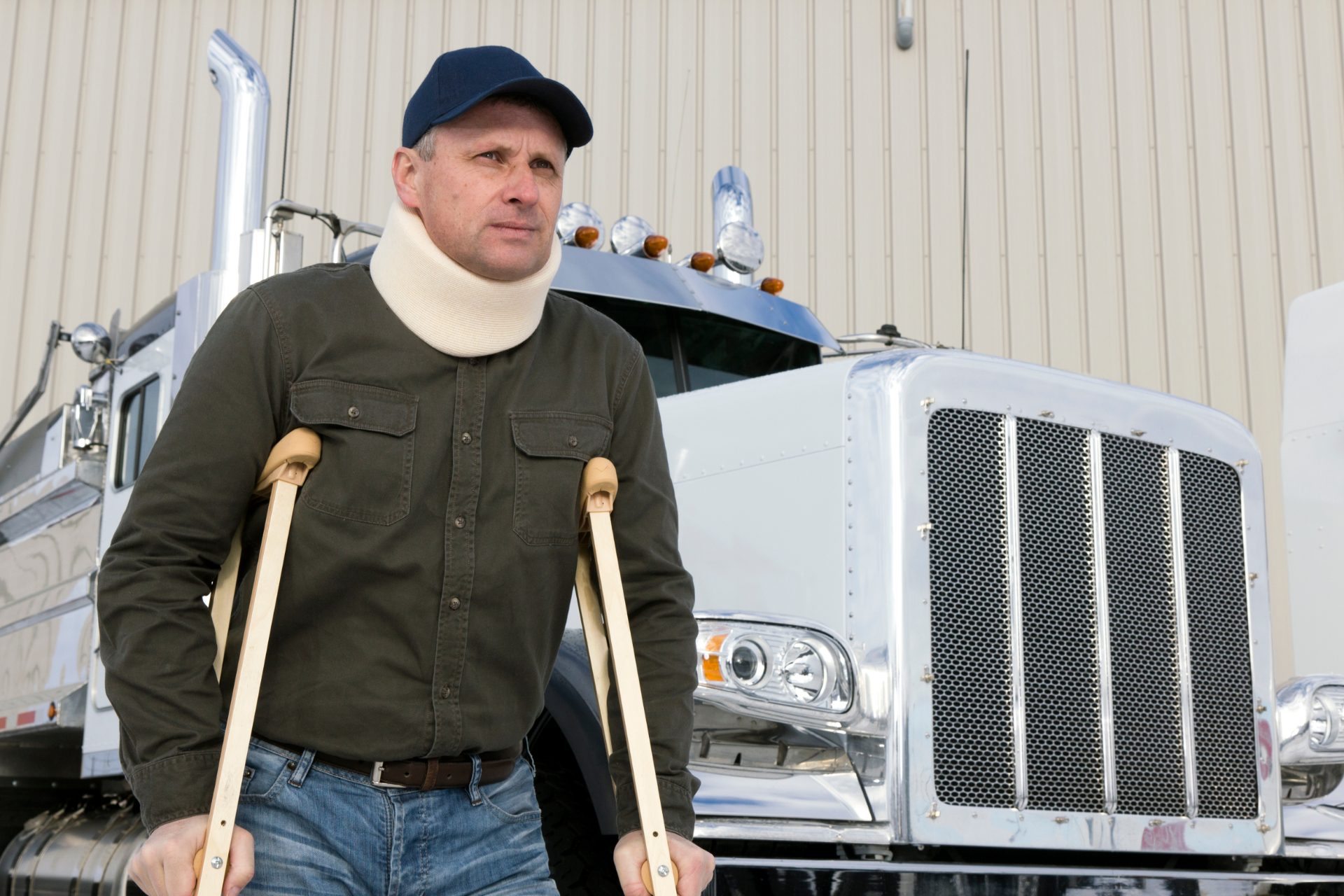 Man standing on crutches in front of a silver semi-truck, wearing a dark cap and jacket