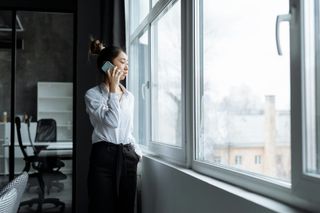 Woman on phone by large windows in an office, looking outside.
