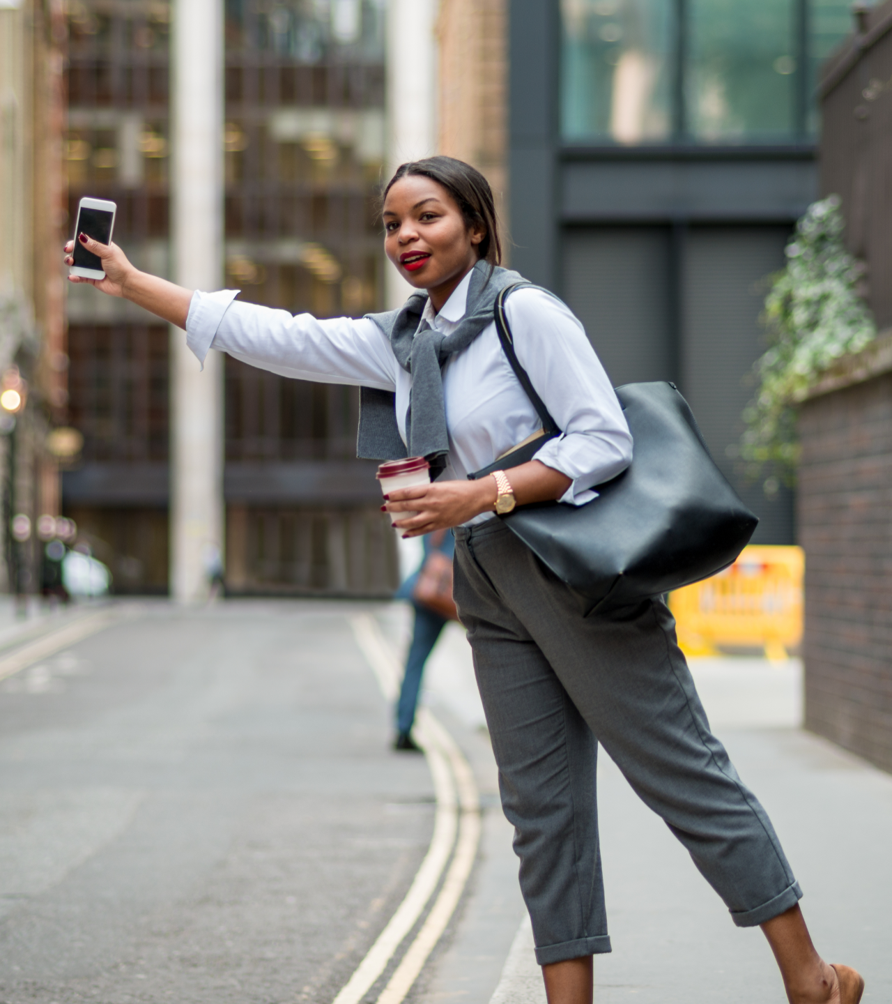 Woman taking a selfie on a city street, holding coffee and a large black bag.