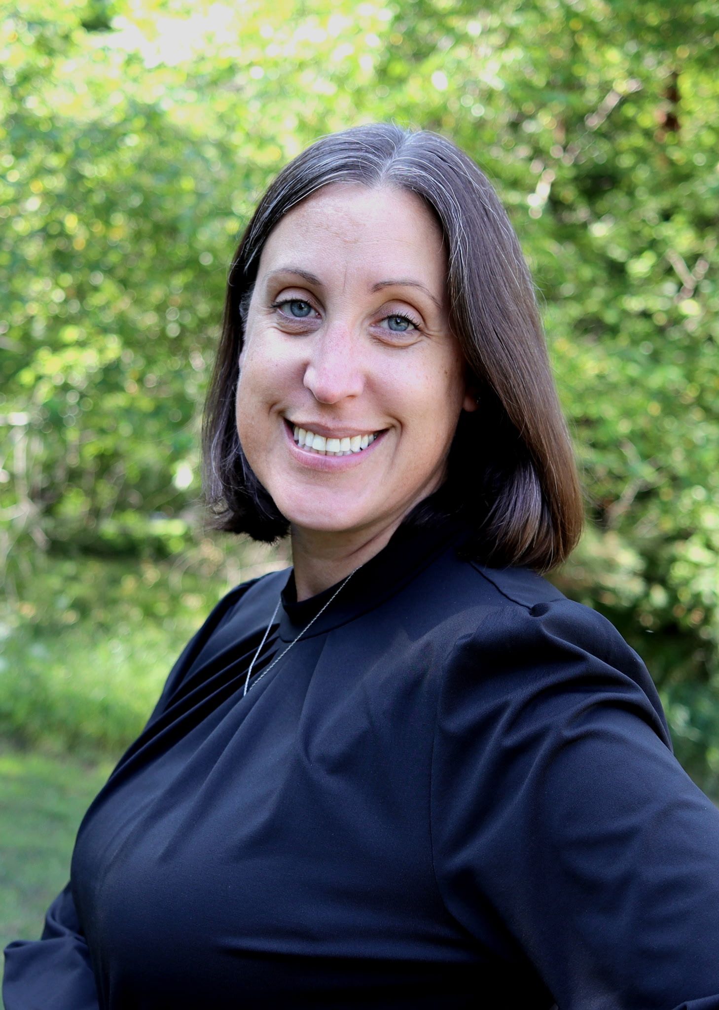 Smiling person in a black shirt outdoors against a leafy green background