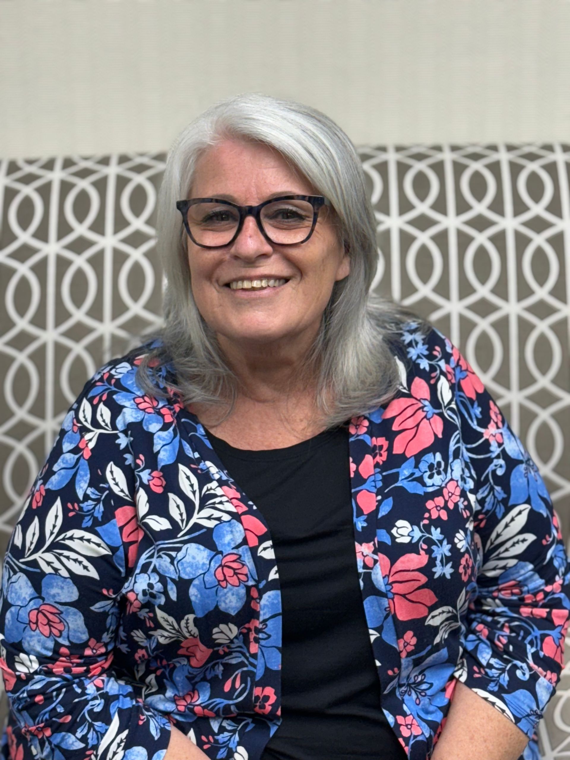 A woman wearing glasses and a floral jacket is smiling