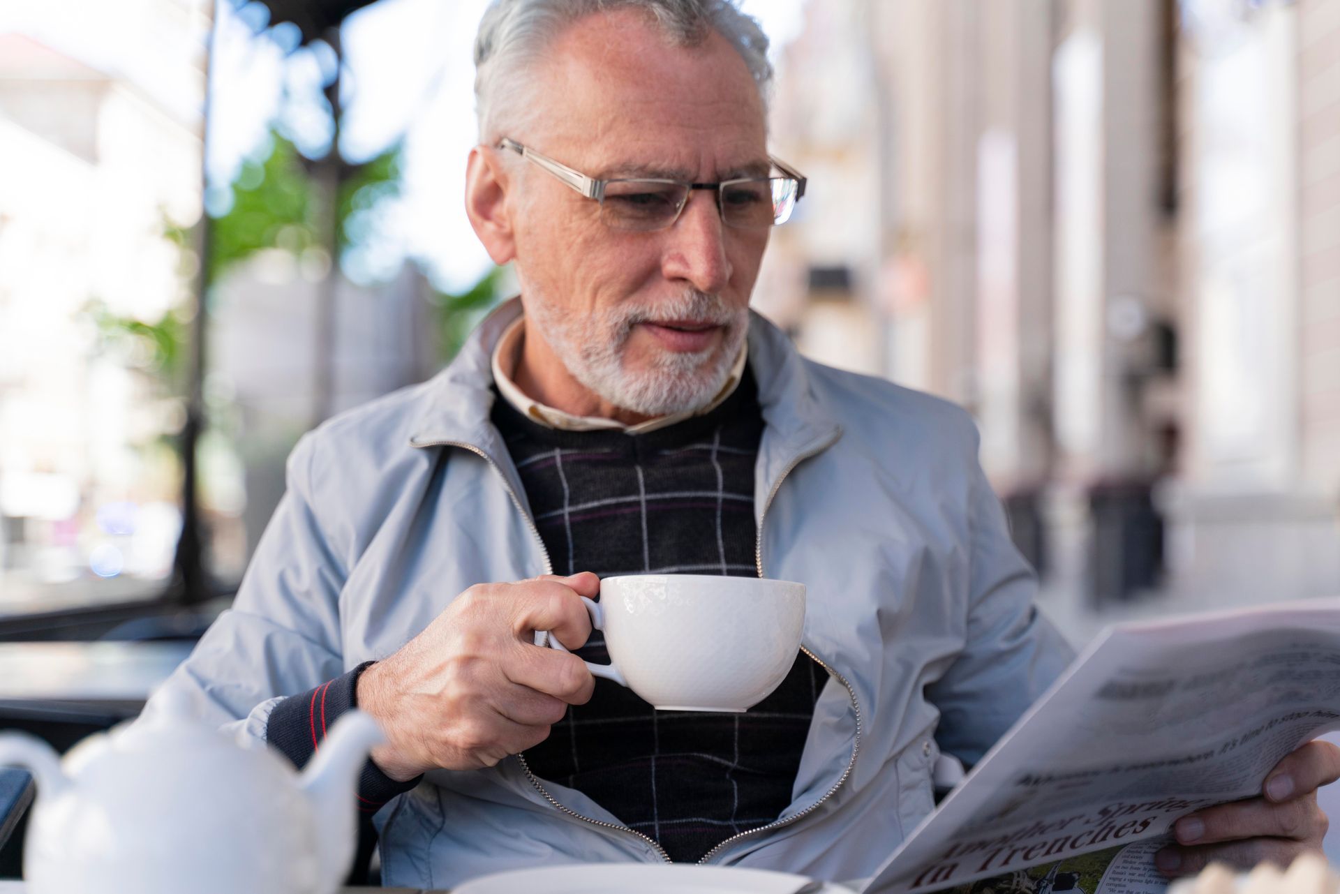 An older man is sitting at a table drinking a cup of coffee and reading a newspaper.