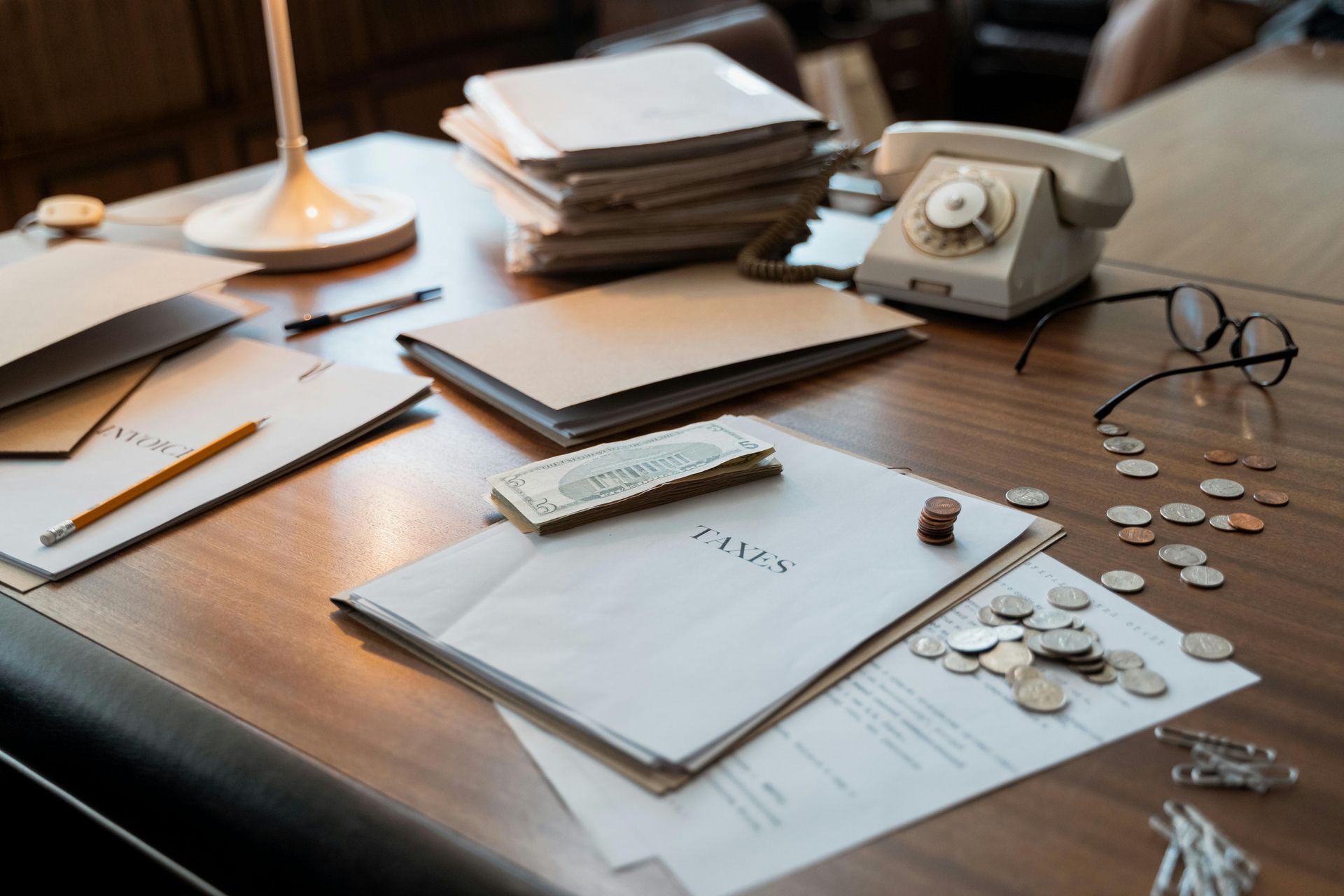 A desk with papers , envelopes , a telephone , glasses and coins on it.