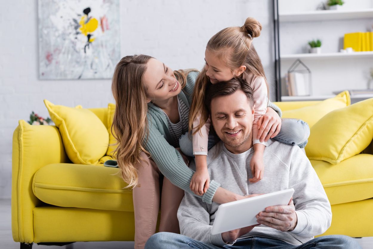 Famiglia seduta su un divano giallo, sorridente e intenta a guardare insieme un tablet in un soggiorno luminoso