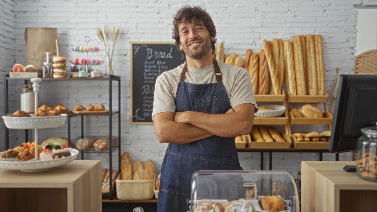 Fornaio in piedi in una panetteria, con le braccia incrociate e un sorriso, dietro i banconi e gli scaffali di pane fresco e pasticcini